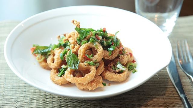 A bowl of battered calamari rings sprinkled with herbs and chili with cutlery and a glass next to it. 